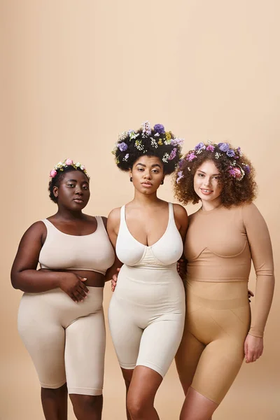 Lächelnde multiethnische Freundinnen in Dessous mit bunten Blumen im Haar auf beige, plus size beauty — Stockfoto
