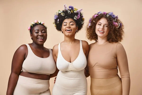 Smiling multiethnic girlfriends in lingerie with colorful flowers in hair on beige, plus size beauty — Stock Photo