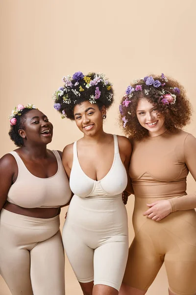 Sonrientes novias multiétnicas en lencería con flores de colores en el pelo en beige, además de belleza de tamaño - foto de stock