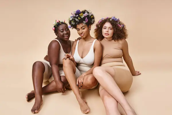 Diverse multiethnic girlfriends in lingerie with flowers in hair sitting on beige, plus size beauty — Stock Photo