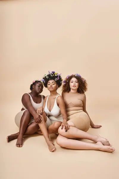 Body positive multiethnische Freundinnen in Dessous mit Blumen im Haar sitzen auf beigem Hintergrund — Stockfoto