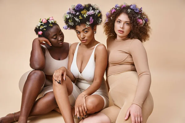 Corps positif femmes multiethniques en lingerie avec des fleurs dans les cheveux assis sur beige, beauté naturelle — Photo de stock