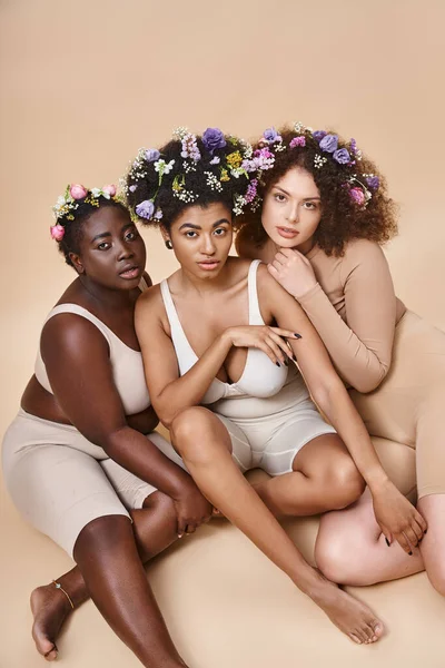 Cuerpo positivo mujeres multiétnicas en lencería con flores en el pelo sentado en beige, belleza natural - foto de stock