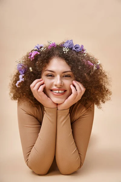 Portrait of pretty and cheerful woman with colorful flowers in wavy hair on beige, natural beauty — Stock Photo
