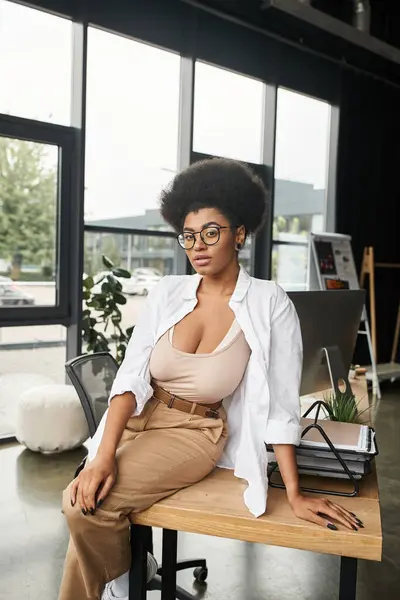 Joven y elegante mujer de negocios afroamericana en gafas sentada en el escritorio de trabajo en la oficina moderna - foto de stock