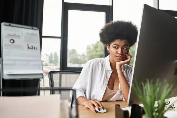 Вдумчивая американская бизнесвумен в очках, работающая за компьютером в современном офисе — стоковое фото