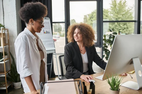 Donna d'affari afroamericana sorridente vicino a collega donna che lavora al computer in ufficio, lavoro di squadra — Foto stock