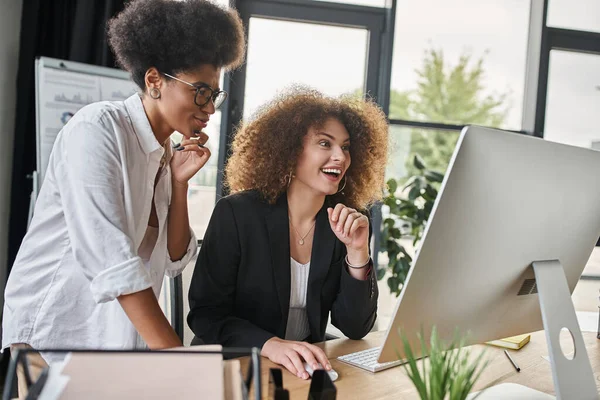 Excited businesswoman looking at computer monitor near african american colleague in modern office — Stock Photo