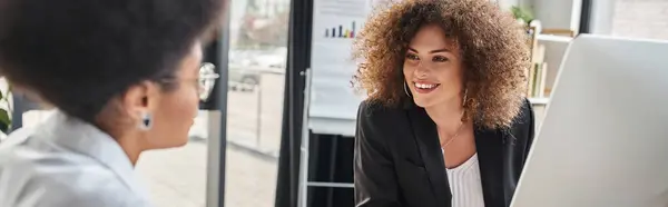 Happy businesswoman talking to african american colleague with notebook in modern office, banner — Stock Photo