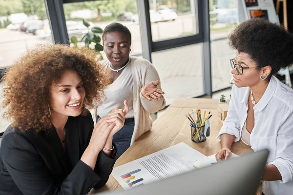 Smiling multiethnic businesswomen looking at computer monitor near documents with graphs in office — Stock Photo