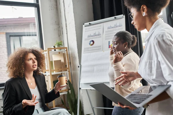 Creative team of multiethnic businesswomen talking near laptop and flip chart in office, startup — Stock Photo