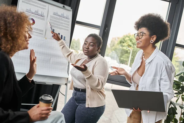 Creative team of multiethnic businesswomen talking near laptop and flip chart in office, startup — Stock Photo