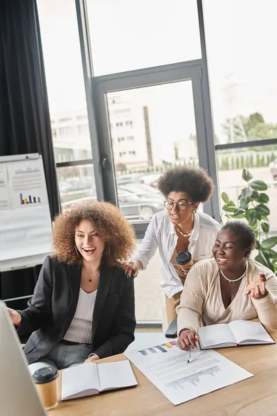 Cheerful multiethnic businesswoman looking at computer monitor near documents with graphs in office — Stock Photo