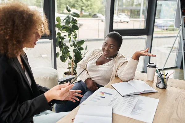 Curvy african american businesswoman talking to colleague near documents with graphs in office — Stock Photo
