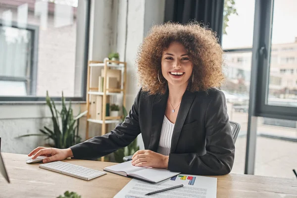 Jovem empresária alegre com cabelo ondulado sentado perto do computador no local de trabalho no escritório moderno — Fotografia de Stock