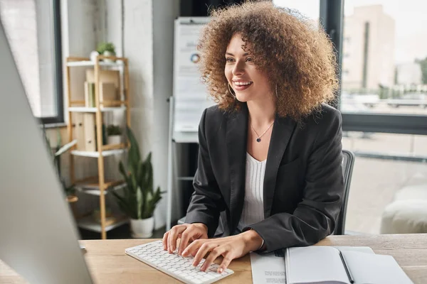 Joven mujer de negocios sonriente con el pelo ondulado escribiendo en la computadora en el lugar de trabajo en la oficina moderna - foto de stock