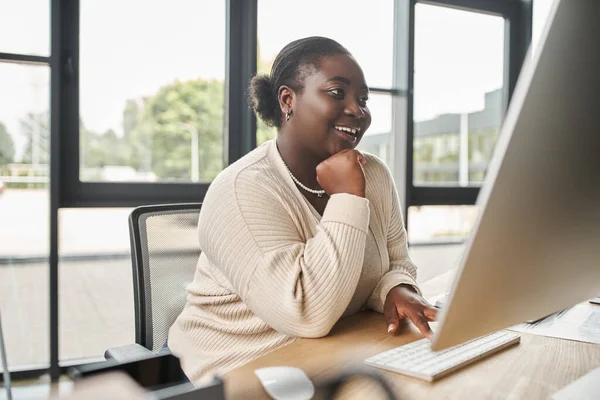 Satisfait afro-américain plus taille femme d'affaires à la recherche sur écran d'ordinateur dans le bureau, le succès — Photo de stock