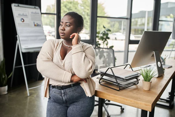 Réfléchie afro-américaine plus taille femme d'affaires détournant les yeux tout en se tenant dans le bureau moderne — Photo de stock