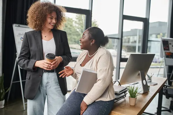 Alegre empresaria multiétnica con café para llevar y carpeta con documentos hablando en la oficina - foto de stock
