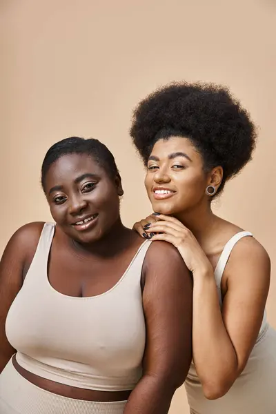 Modelos femininos afro-americanos alegres em roupa interior olhando para a câmera em bege, além de beleza tamanho — Fotografia de Stock