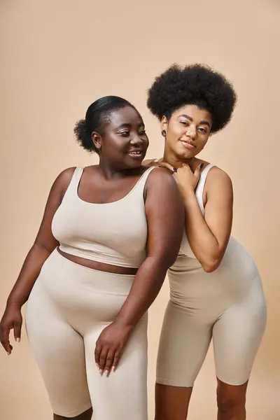 Modelos femeninos afroamericanos alegres en ropa interior posando en beige, belleza natural más tamaño - foto de stock