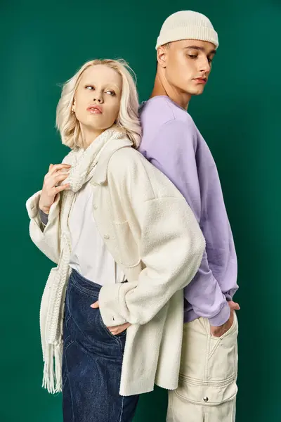 Блондинка в зимнем наряде позирует с рукой в кармане и спиной к спине с высоким мужчиной на бирюзовом — стоковое фото