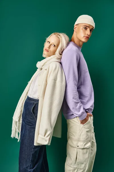 Blonde Frau in Winterkleidung steht Rücken an Rücken mit jungem Mann in Mütze vor türkisfarbener Kulisse — Stockfoto