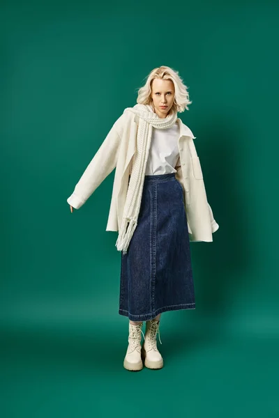 Стильная блондинка в белой куртке и джинсовой юбке позирует на бирюзовом фоне, зимняя мода — стоковое фото