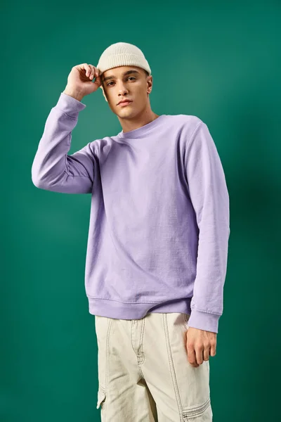 Handsome man in purple sweatshirt adjusting beanie hat on turquoise backdrop, winter fashion — Stock Photo