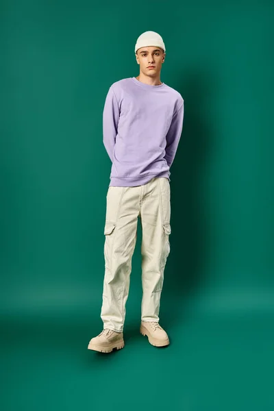 Longueur totale de l'homme en sweat-shirt violet et chapeau de bonnet posant sur fond turquoise, mode d'hiver — Photo de stock