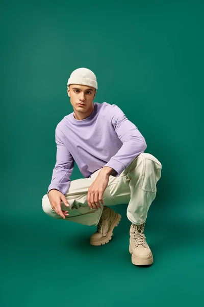 Full length of man in purple sweatshirt and beanie hat sitting on turquoise backdrop, winter fashion — Stock Photo