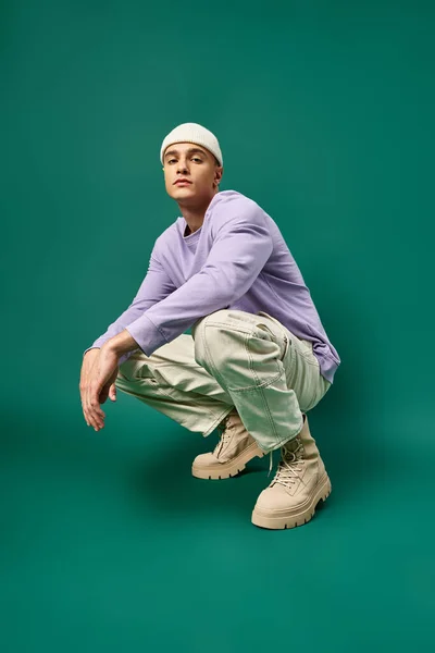 Full length of man in purple sweatshirt and beanie hat sitting on turquoise backdrop, winter style — Stock Photo