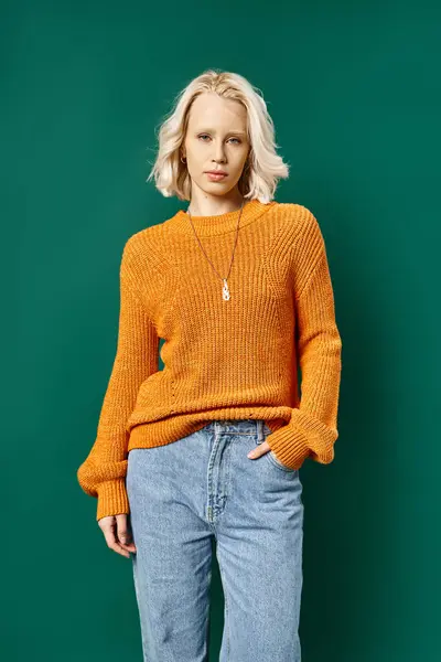 Красива блондинка в гірчичному жовтому светрі і джинсах позує з рукою в кишені на бірюзовому — стокове фото