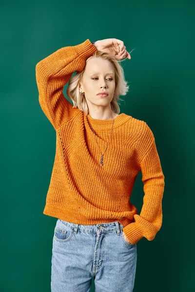 Молода блондинка в гірчичному жовтому светрі і джинсах позує з рукою в кишені на бірюзовому — стокове фото