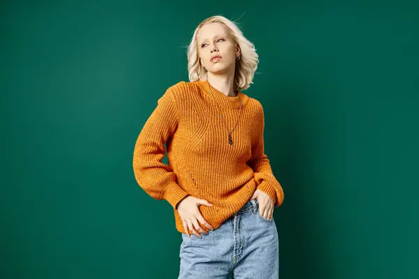 Blonde woman in mustard yellow sweater posing with hand in pocket of jeans on turquoise backdrop — Stock Photo