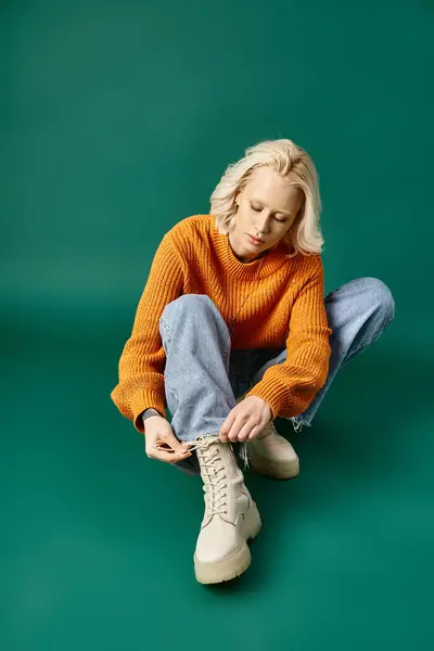 Tattooed woman in mustard yellow sweater and jeans tying laces on boots on turquoise background — Stock Photo