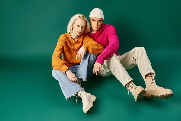 Elegante pareja en suéteres, mujer rubia y hombre en gorro sentado juntos sobre fondo turquesa - foto de stock