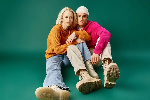 Pareja de moda en suéteres, mujer rubia y hombre en gorro sentado juntos sobre fondo turquesa - foto de stock