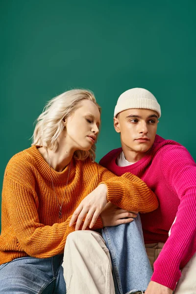 Stylish couple in sweaters, blonde woman and man in beanie sitting together on turquoise background — Stock Photo