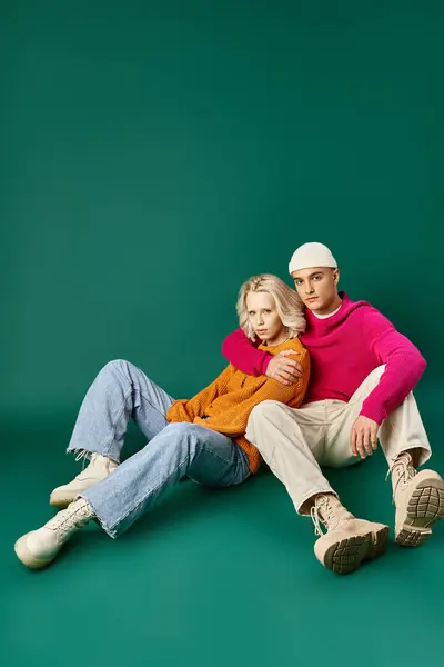 Elegante pareja en suéteres, hombre en gorro abrazando a la mujer rubia mientras están sentados juntos en turquesa - foto de stock