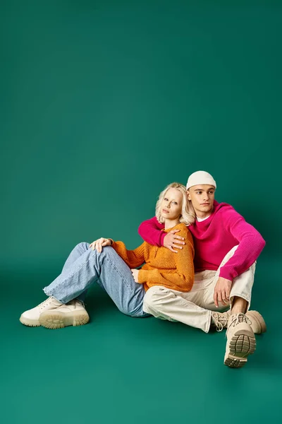Couple en pulls, beau homme en bonnet embrassant femme blonde tout en étant assis ensemble sur turquoise — Photo de stock