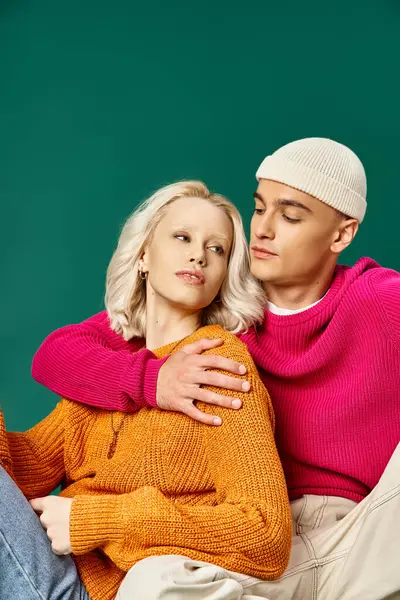 Stylish couple in sweaters, man in beanie hugging blonde woman while sitting together on turquoise — Stock Photo