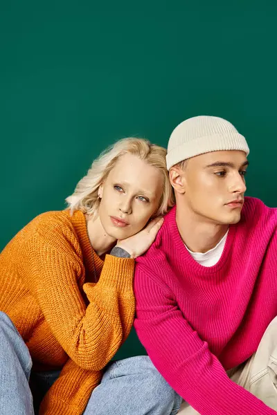 Stylish couple in sweaters, dreamy woman leaning on shoulder of man in beanie, sitting on turquoise — Stock Photo