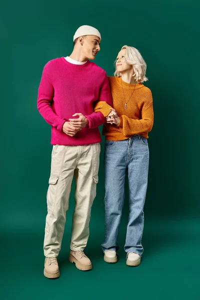 Feliz pareja en suéteres de invierno de pie mano a mano sobre fondo turquesa, mujer joven y hombre - foto de stock