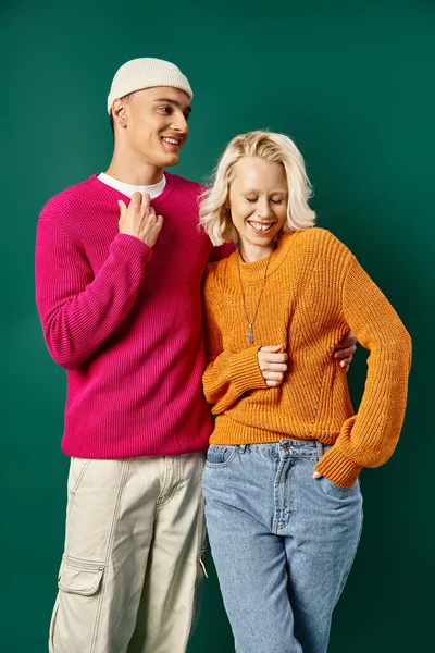 Cheerful young couple in winter attire laughing and standing together on turquoise background — Stock Photo