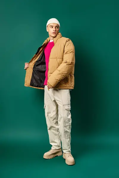 Молодой человек в шапке шапочки и стильной куртке фугу позировать на бирюзовом фоне, зимняя мода — стоковое фото