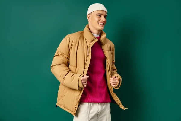 Весёлый молодой человек в шапочке и стильной куртке на бирюзовом фоне, зимние тренды — стоковое фото