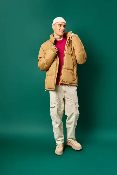 Hombre joven feliz en gorro sombrero y chaqueta hinchable con estilo sobre fondo turquesa, tendencias de invierno - foto de stock