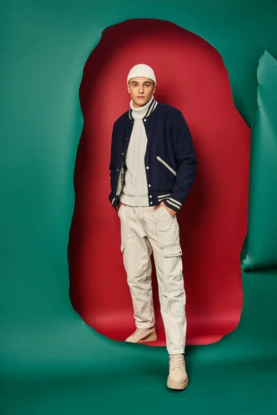 Стильный молодой человек в шапочке, белый свитер и куртка бомбардировщика на разорванном красном с бирюзовым фоном — стоковое фото