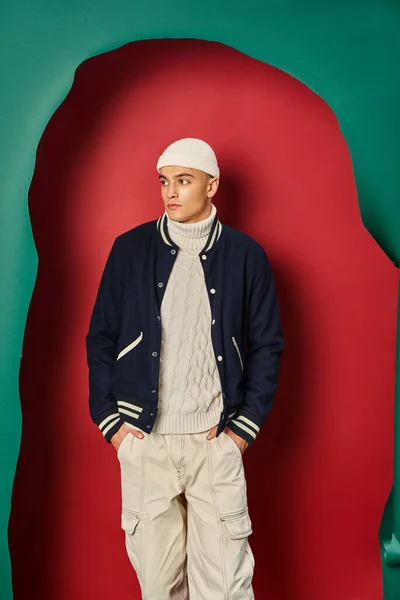 Jeune homme en bonnet, pull blanc et veste de bombardier posant sur rouge déchiré avec fond turquoise — Photo de stock